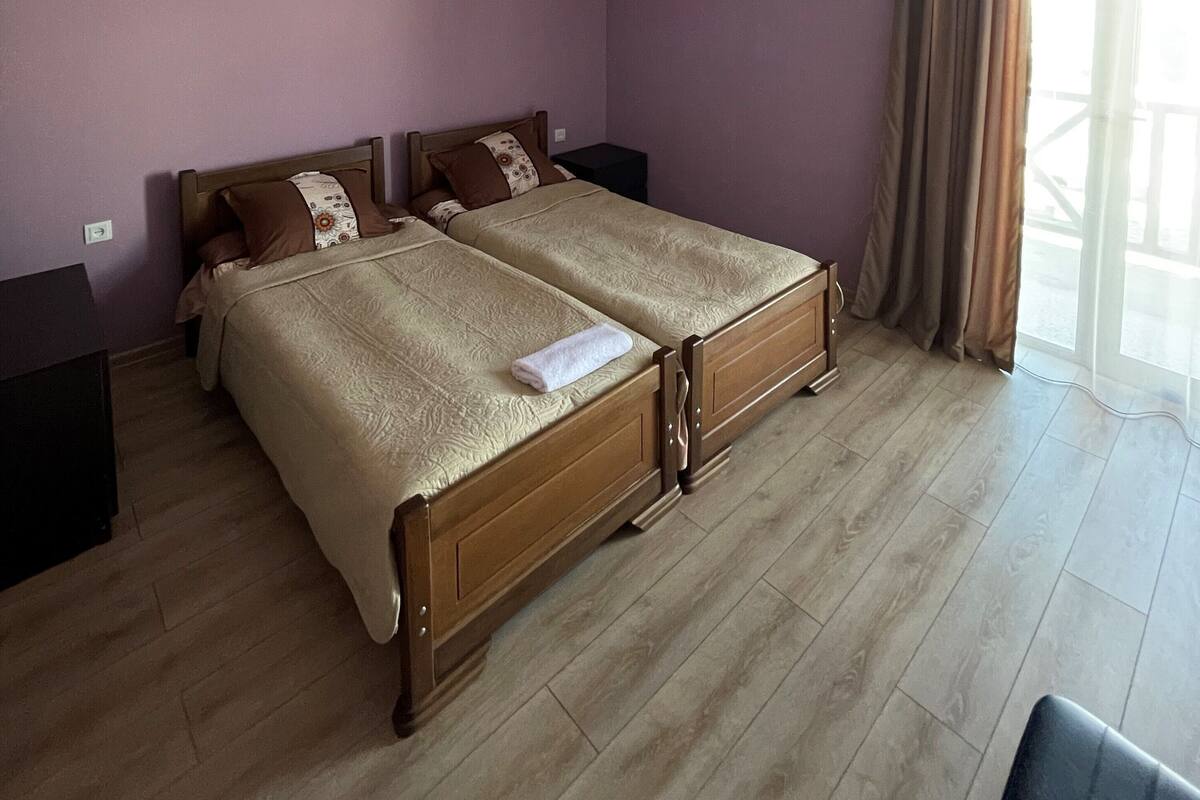 A double bed with a wooden frame and two pillows, a bedside table, and a window with curtains.