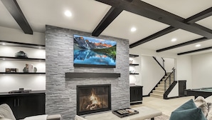 A modern living room with a stone fireplace, a flat-screen TV mounted above it, and a cozy seating area with a sofa and cushions.