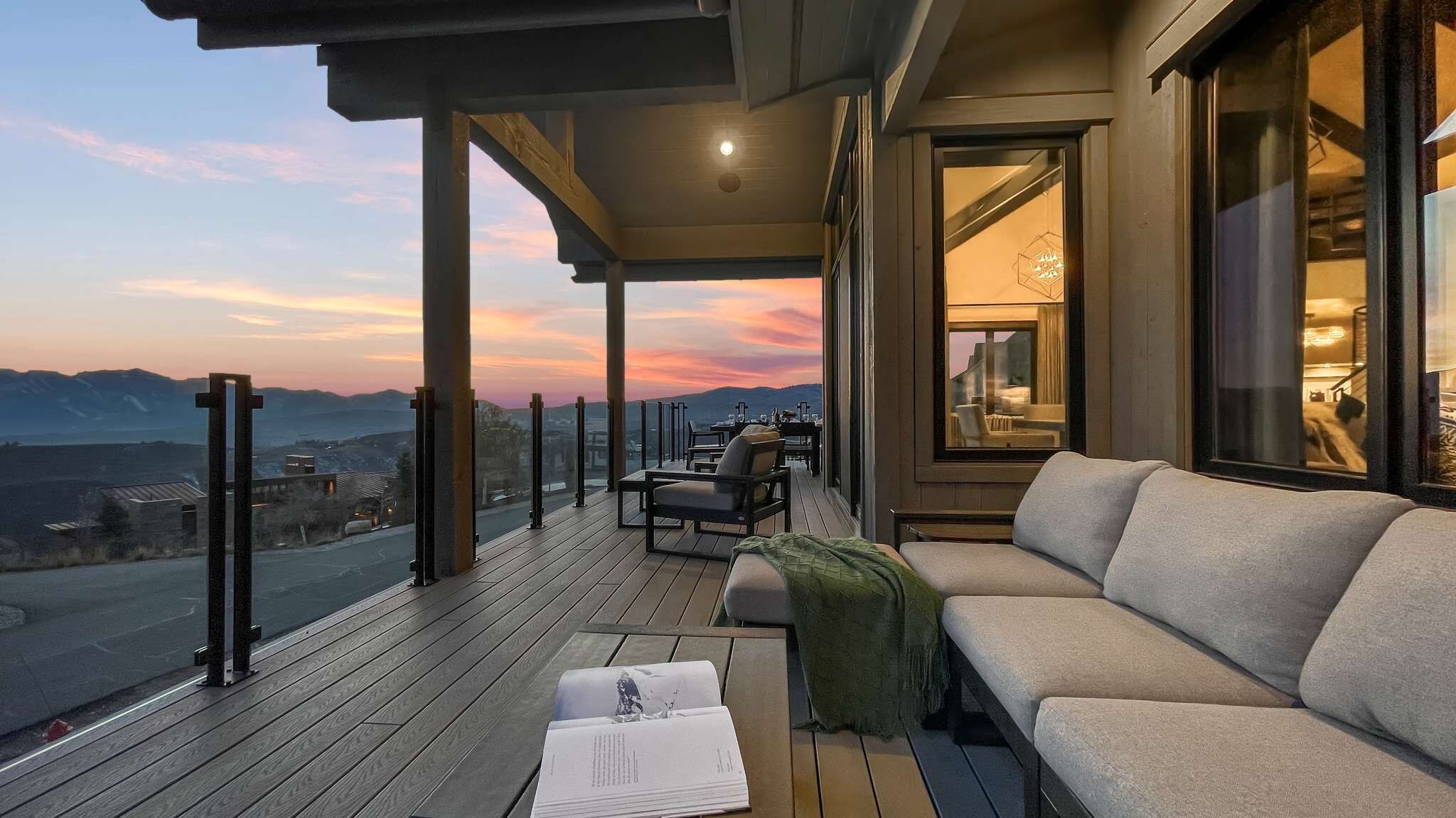 Un balcón con sofá, una mesa y un libro, con vistas a un paisaje con montañas y un atardecer.