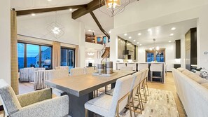 A spacious dining area with a long table and multiple chairs, a kitchen with modern appliances, and a view of the outside through large windows.