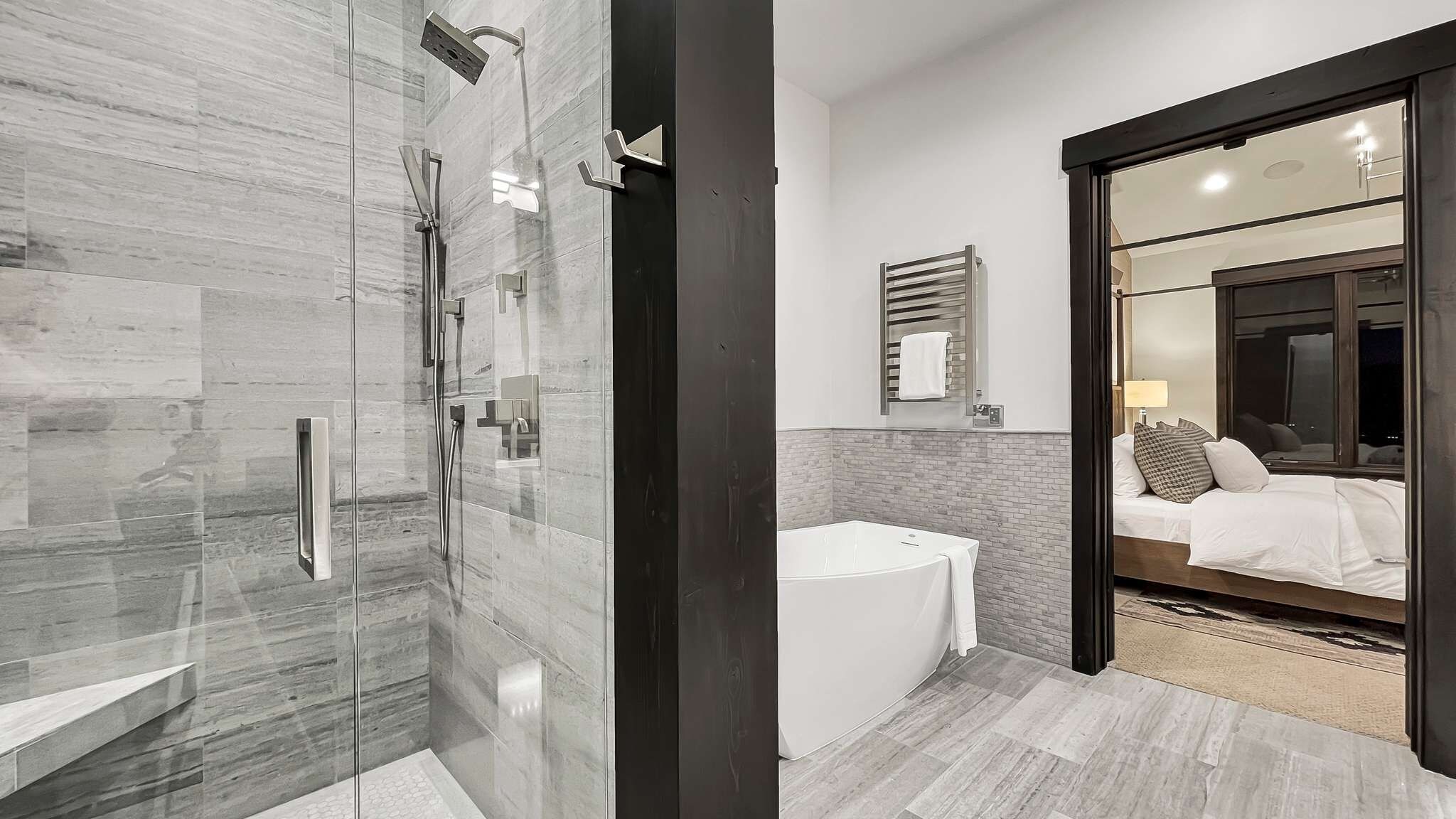 A modern bathroom with a glass-enclosed shower, a white sink, and a large mirror reflecting a bedroom with a bed and a nightstand.