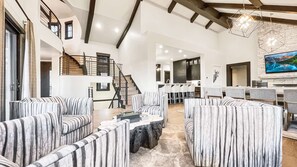 A spacious living area with striped sofas, a staircase, a kitchen, and a large TV screen.