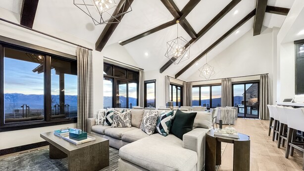 A spacious living room with a large sofa, a coffee table, and a dining area with a table and chairs. Large windows offer a view of the mountains.