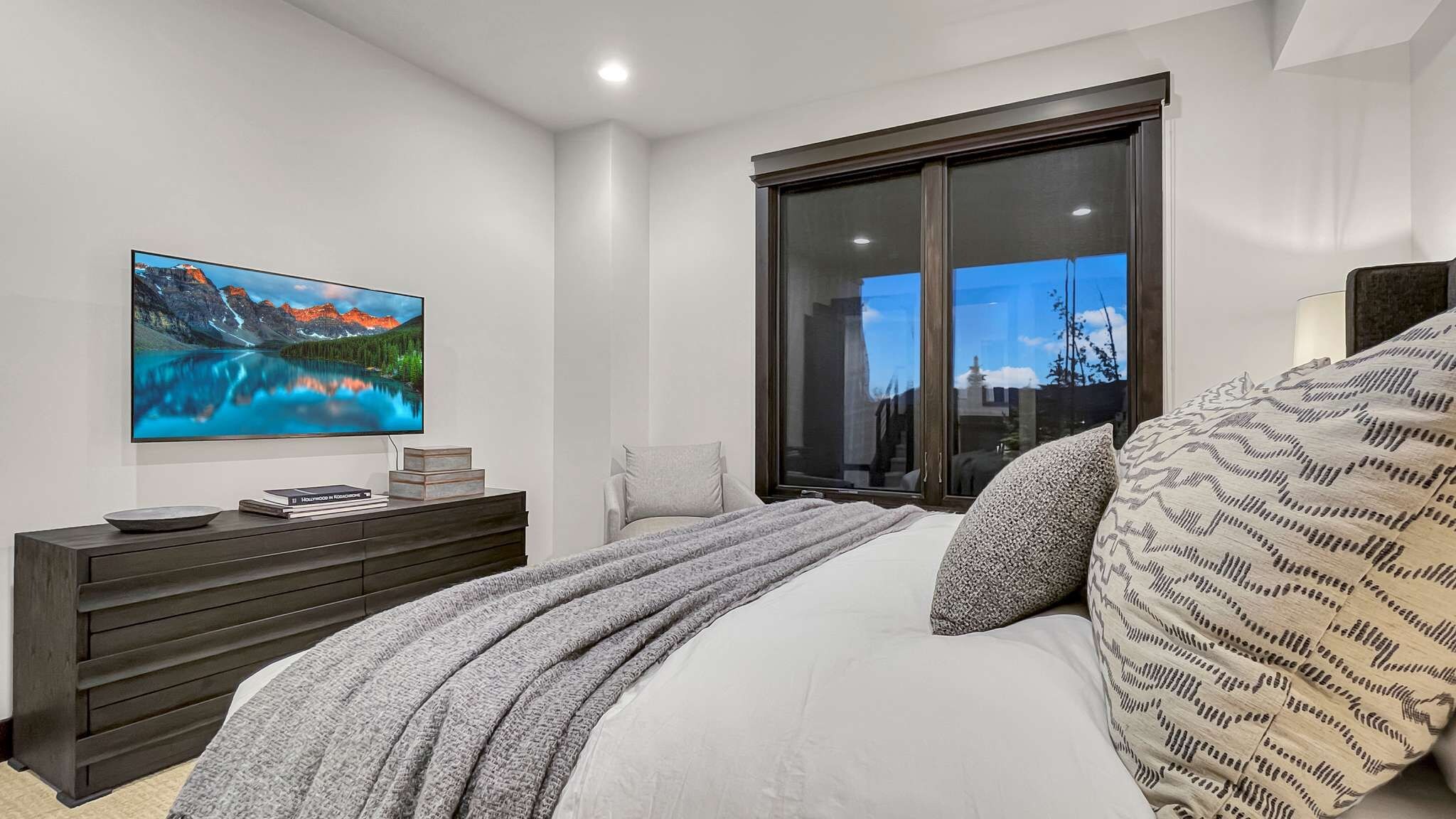 Een slaapkamer met een groot bed, een flatscreen televisie aan de muur, een donker houten dressoir en een groot raam met uitzicht op de stad.