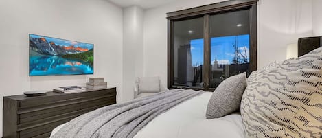 A bedroom with a large bed, a flat-screen TV mounted on the wall, a dark wooden dresser, and a large window with a view of the city.