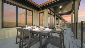 Un área de comedor al aire libre con una mesa lista para dos personas, rodeada de sillas y grandes ventanas que ofrecen vistas del paisaje.