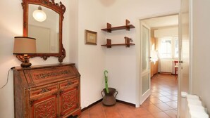 Um corredor com um armário de madeira, um espelho e um guarda-chuva verde em vaso. Há prateleiras fixadas na parede e uma porta que dá para outro quarto.