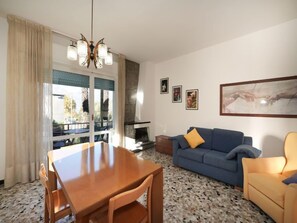 A living room with a wooden dining table, blue sofa, and yellow armchair. There is a fireplace, a chandelier, and artwork on the walls.