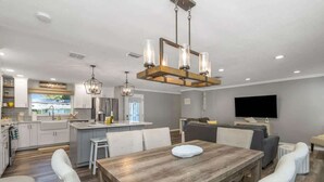 A modern kitchen with white cabinets, a dining area with a wooden table and white chairs, and a living area with a gray sofa and a flat-screen TV.