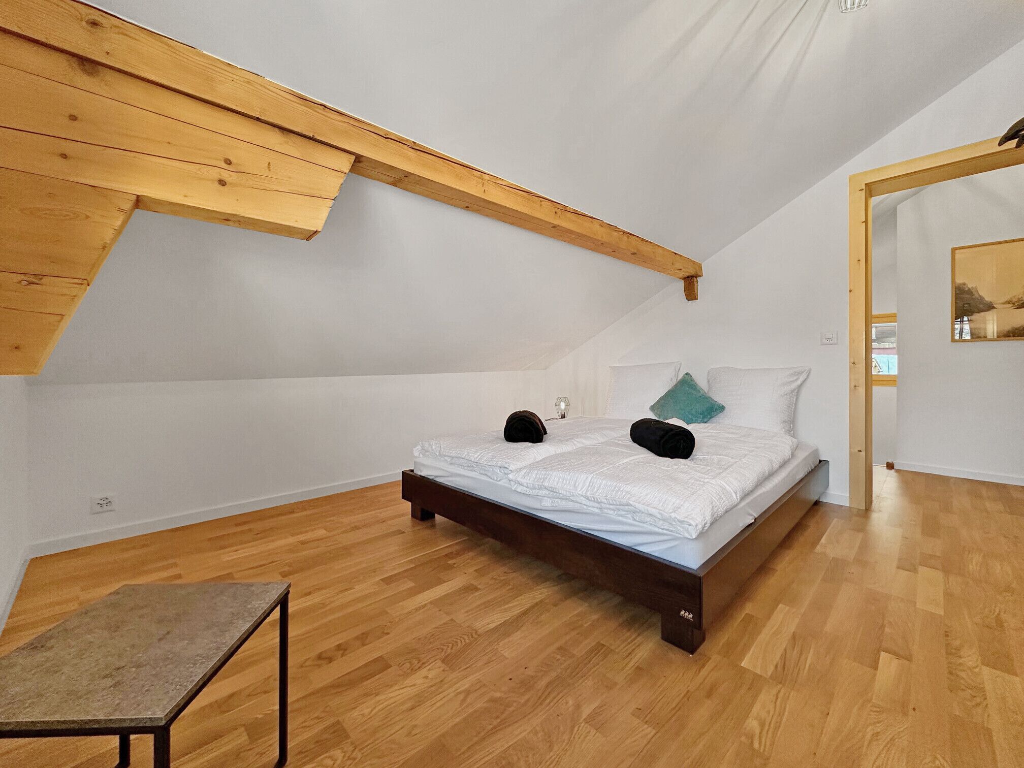 Een slaapkamer met een schuine balkenplafond, een bed met wit beddengoed, een klein tafeltje en een spiegel aan de muur.