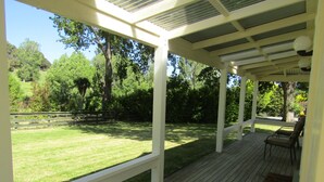 Un patio techado con piso de madera, barandas blancas y vista a un jardín cubierto de césped y árboles.