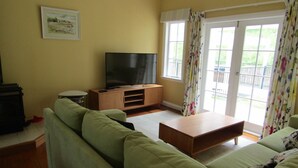 Una sala de estar con un sofá verde, una mesa de centro de madera y un televisor de pantalla plana sobre un mueble de madera. Se ve una ventana con cortinas florales y un balcón a través de las puertas de vidrio.