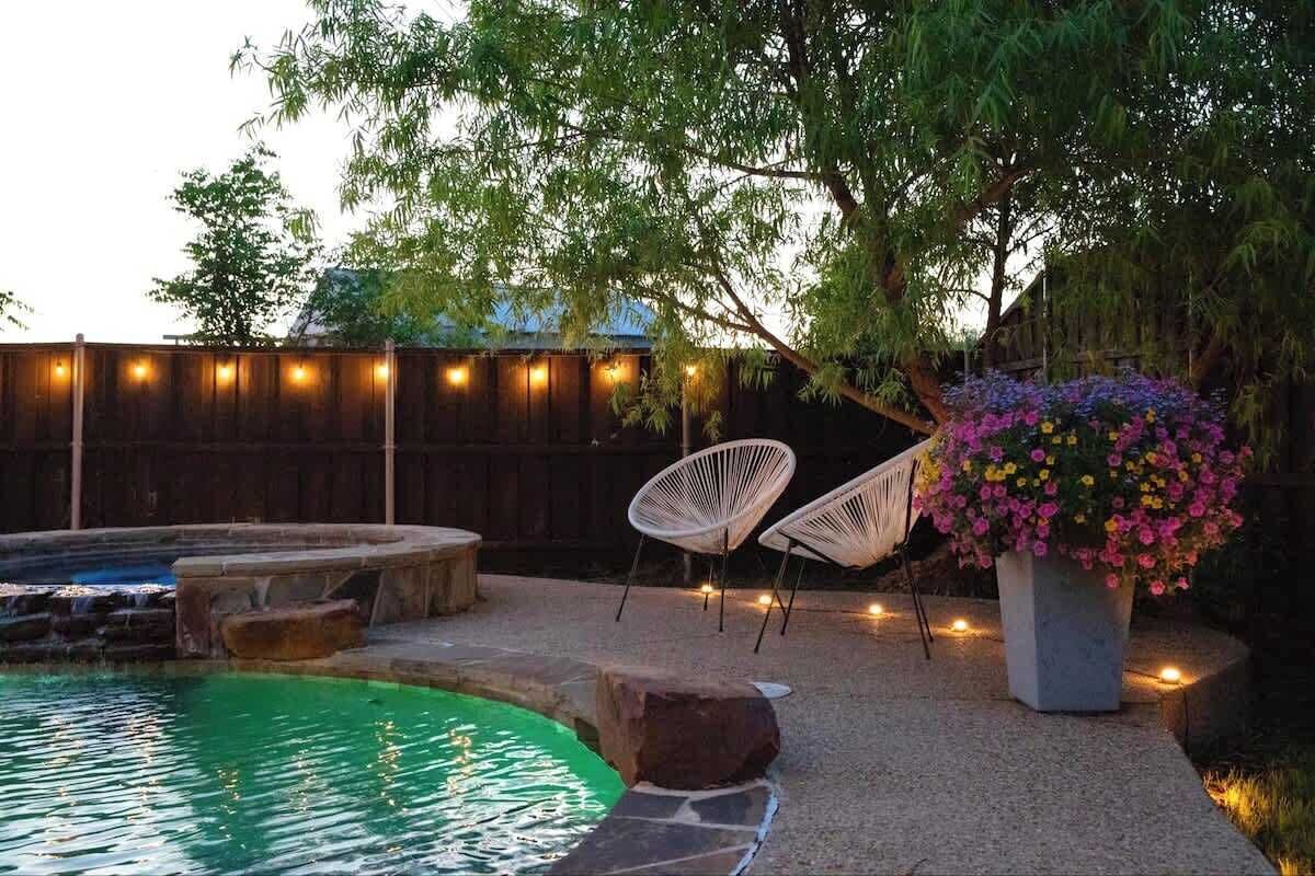 A backyard with a swimming pool, a hot tub, and a wooden fence. There are two white chairs and a large planter with flowers.