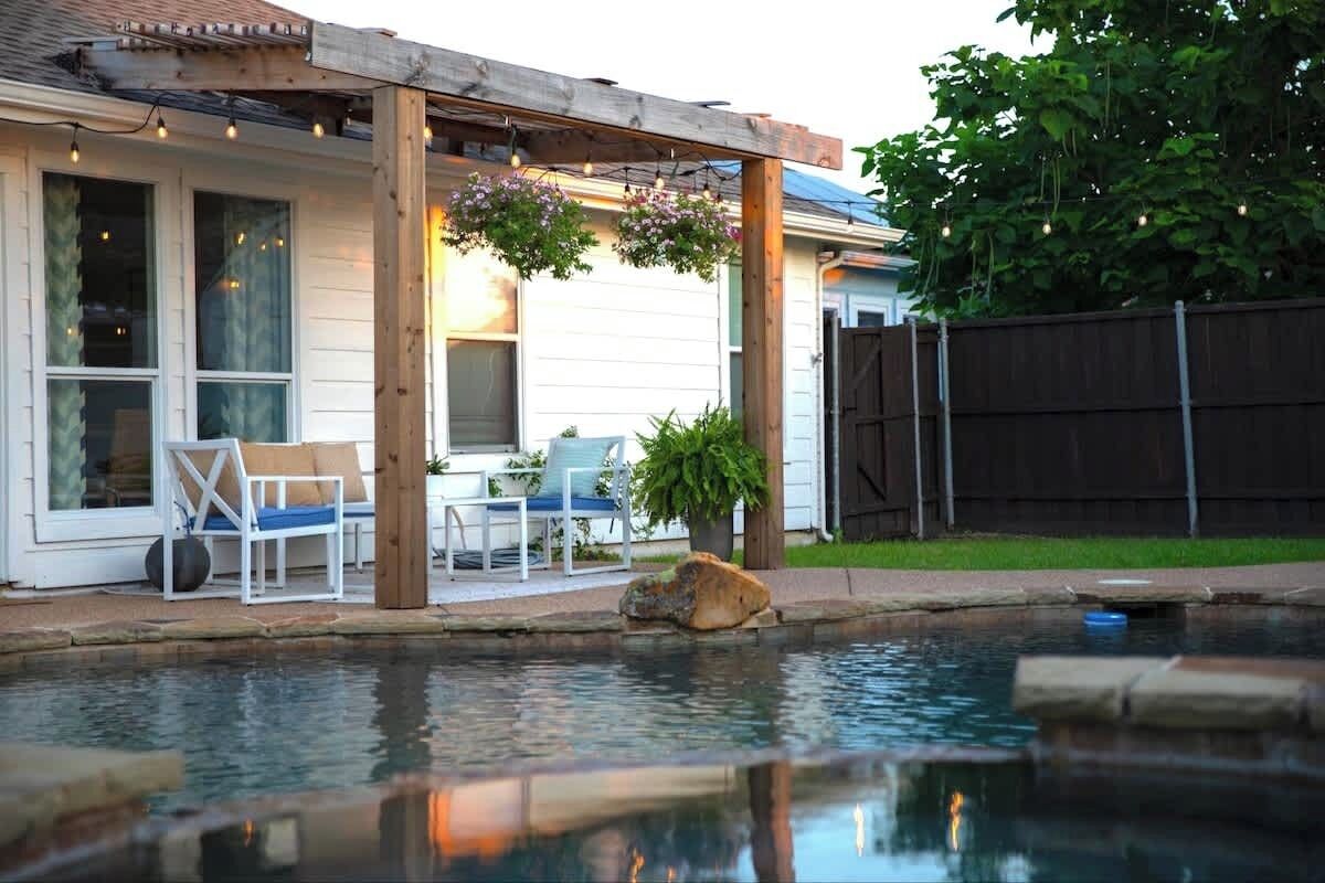 A backyard with a pool, wooden pergola, outdoor furniture, and a fence.