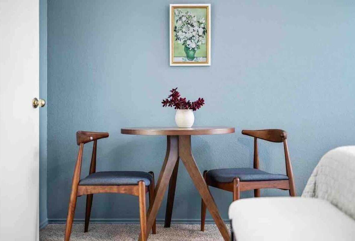 A room with a blue wall, a round wooden table with two chairs, and a vase with flowers.