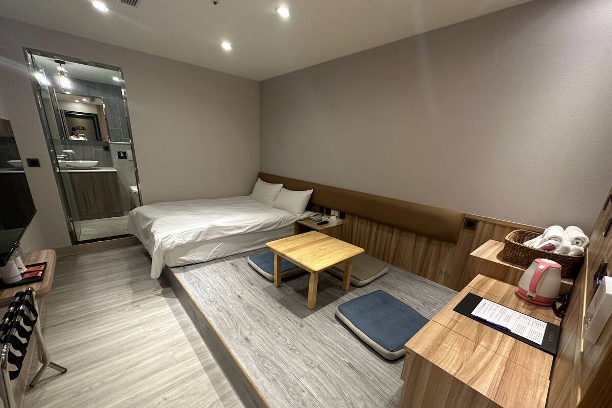 A hotel room with a bed, a wooden table, a small desk, a basket of towels, and a kettle.