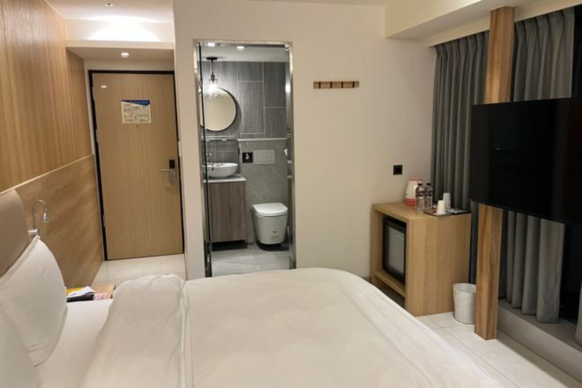 Habitación de hotel con cama, televisión, baño con lavamanos e inodoro, y un mueble de madera con artículos de aseo.