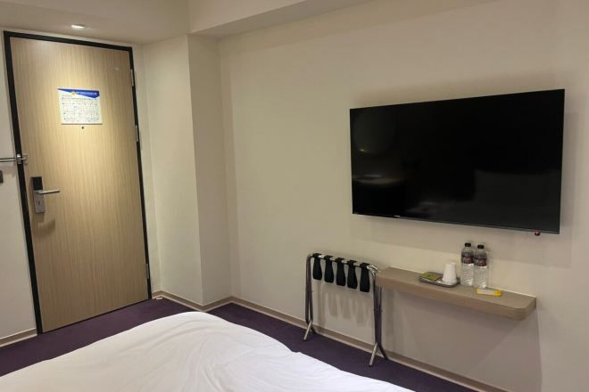 Habitación de hotel con cama, televisor montado en la pared, radiador y un estante pequeño con botellas y una taza.