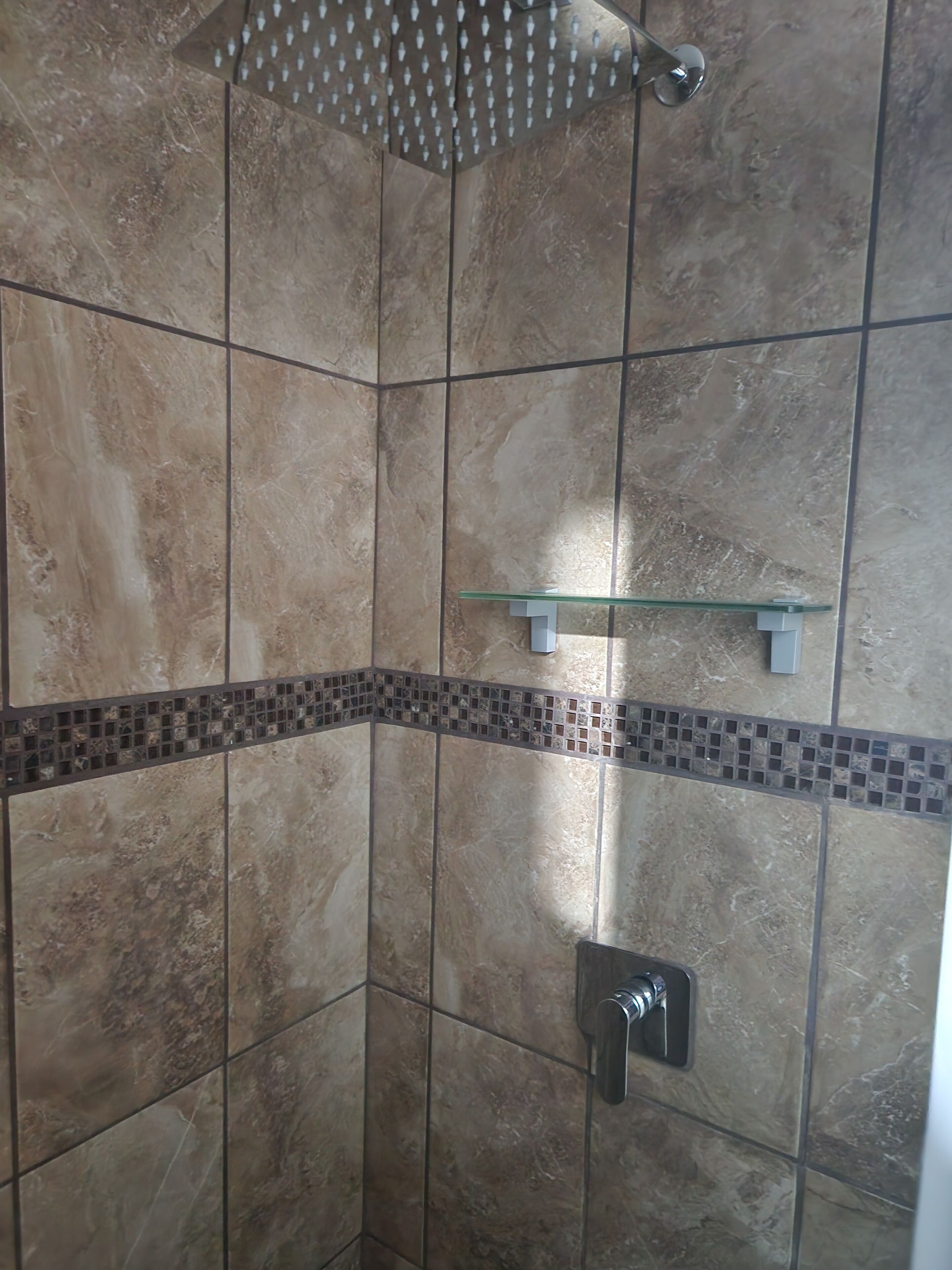 Un espacio de ducha con ducha de lluvia, estante de vidrio y pared con azulejos.