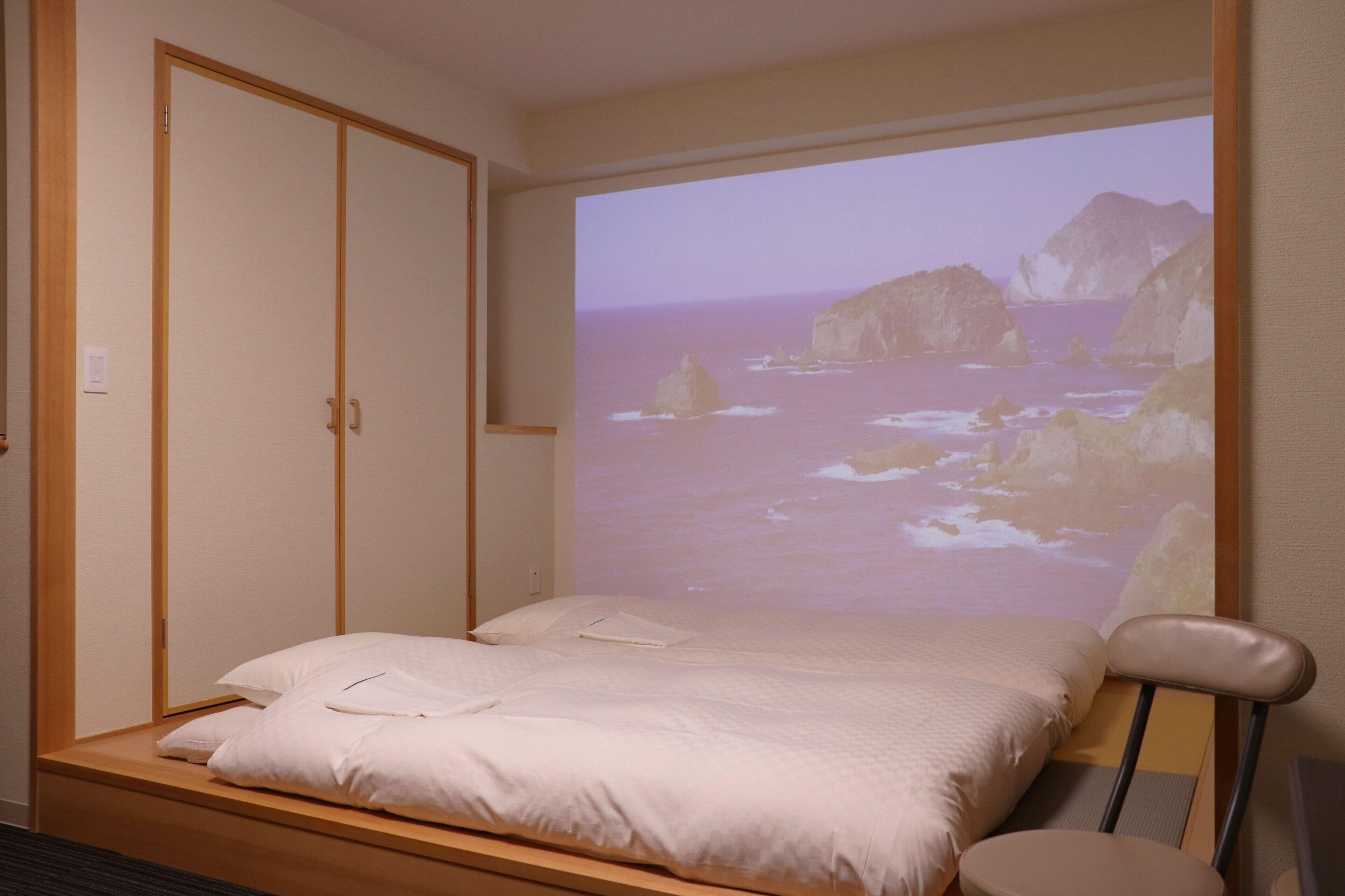 Un dormitorio con una cama, un armario de madera y una pantalla de proyección que muestra una escena costera.