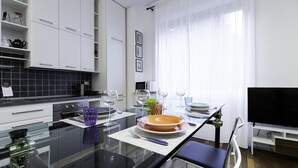 A modern kitchen with white cabinets, a black tiled backsplash, a glass dining table, and a flat-screen TV. - Italianway Easy - Piceno 44 (Milan)