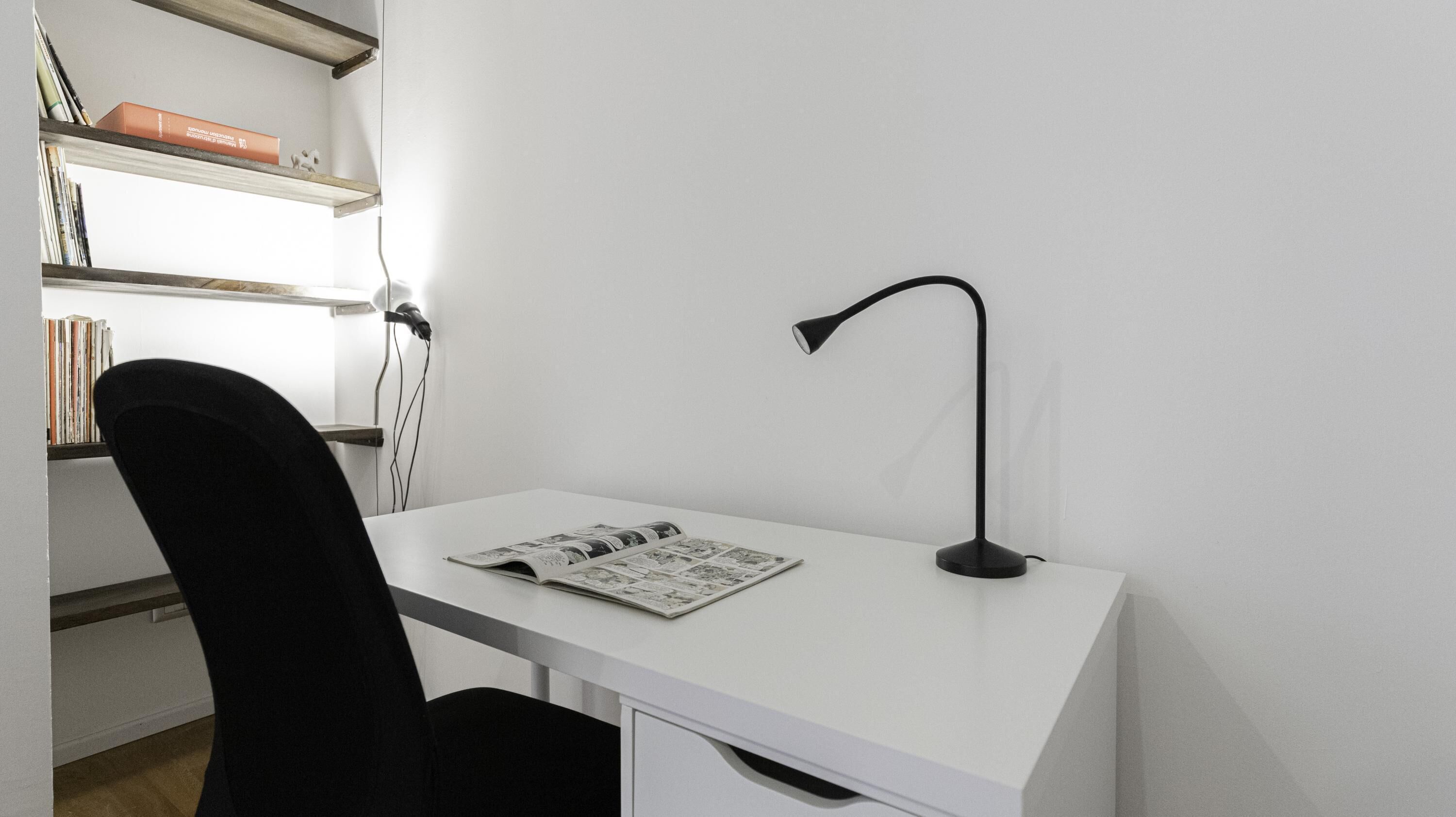 Ein weißer Schreibtisch mit einer schwarzen Schreibtischlampe, einem schwarzen Stuhl und einer Zeitschrift darauf.