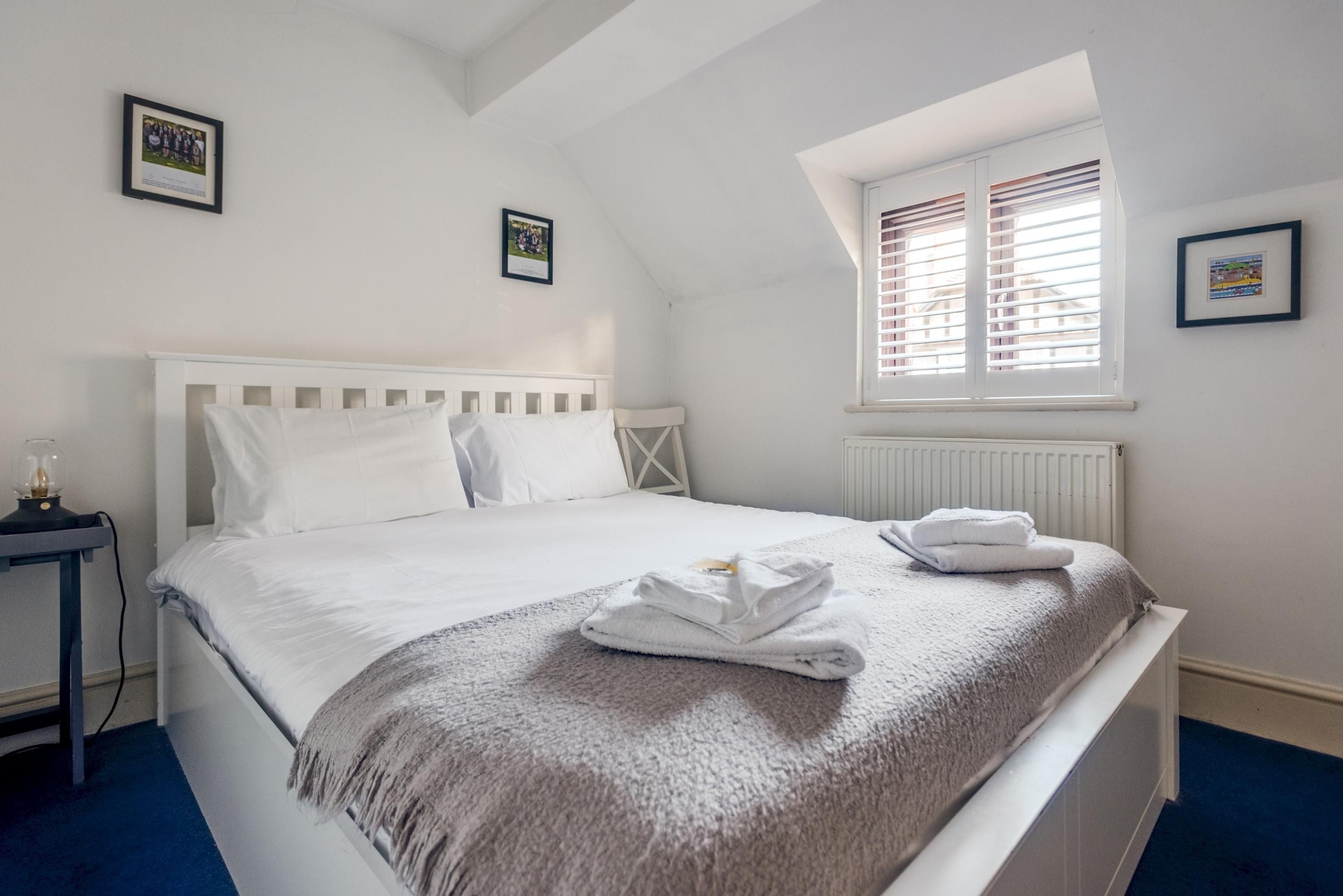 En pænt redt seng med hvidgavl, grå tæppe og to foldede håndklæder. Et lille bord med lampe står ved siden af sengen, og der er et vindue med persienner på væggen.