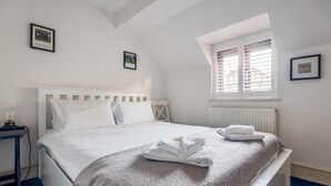 A neatly made bed with a white headboard, a grey throw blanket, and two folded towels on the bed. There is a small table with a lamp beside the bed, and a window with blinds on the wall. - Cosy 2-bedroom Haven in Picturesque Headington (Oxford)