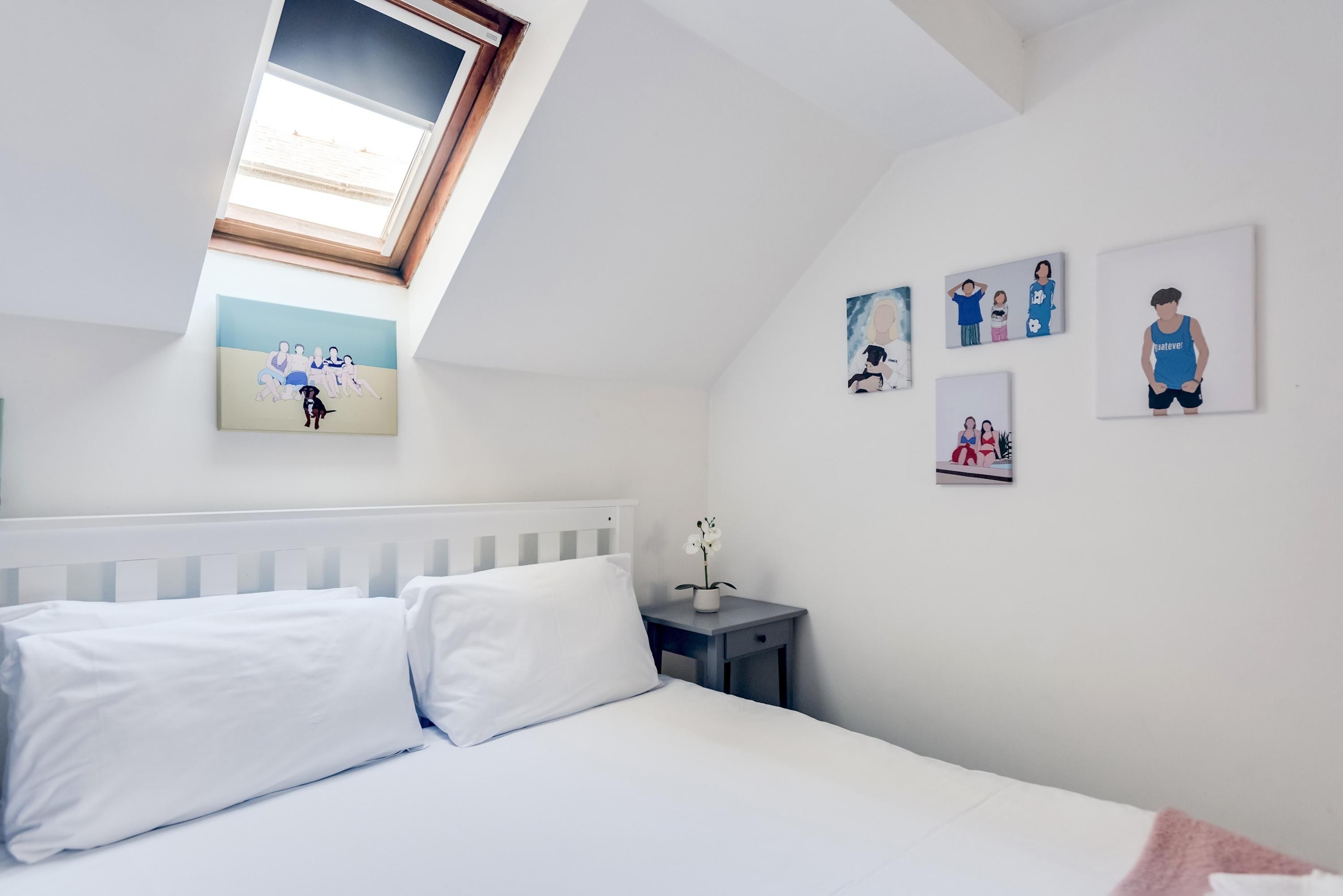 Ein Schlafzimmer mit schrägen Wänden, einem Bett mit weißen Bettwäsche, einem kleinen Nachttisch mit Blumenvase und Wandbildern, die Personen und einen Hund zeigen.