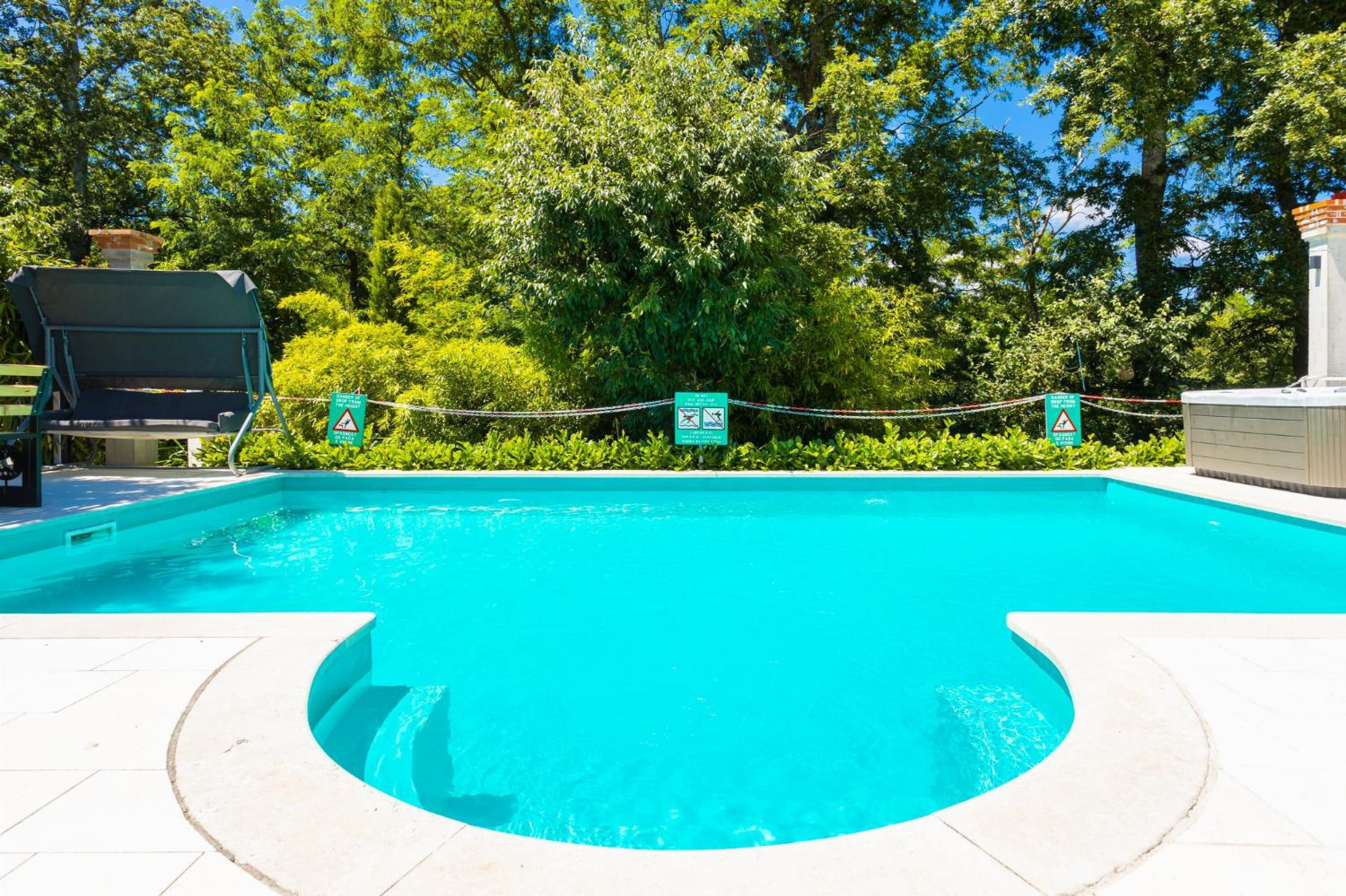 Una piscina amb vora corba, envoltada per un petiter i arbres. Hi ha senyals que indiquen prohibició de salts i de bades.