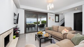 A modern living room with a sofa, coffee table, and fireplace. There is a sliding glass door leading to an outdoor area with a pool and dining table. - Villa Metis in Rhodes (Rhodes)