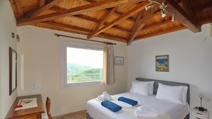 Quarto com teto de madeira, cama com roupa de cama branca, janela com vista, mesa de cabeceira e escrivaninha com cadeira.