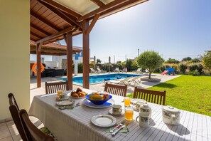 A dining table set outdoors with a pool view, surrounded by chairs and a covered area. - Hermione Villa in Gennadi (Rhodes)