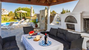 A covered outdoor seating area with a table set for dining, lounge chairs, and a view of a pool and buildings in the background.
