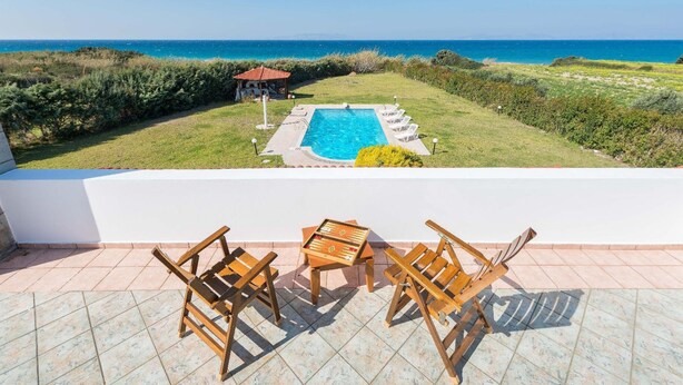A pool with a view of the sea, a gazebo, and wooden chairs on a tiled patio.