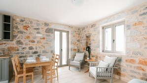A room with stone walls, a wooden dining table with chairs, a white armchair, a small round table with a plant, and a window. - Villa Dioni Ena in Ditiki Mani (West Mani)