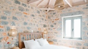 A bedroom with a stone wall, wooden ceiling beams, a bed, a nightstand, a lamp, and a window. - Villa Dioni Ena in Ditiki Mani (West Mani)