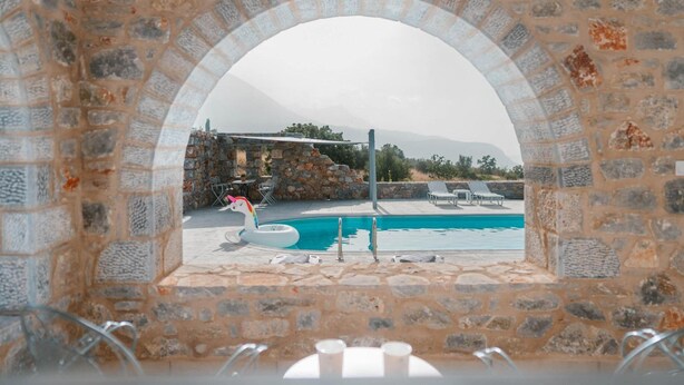 A stone archway leading to a pool area with a stone wall, lounge chairs, and a unicorn float. - Villa Dioni Ena in Ditiki Mani (West Mani)