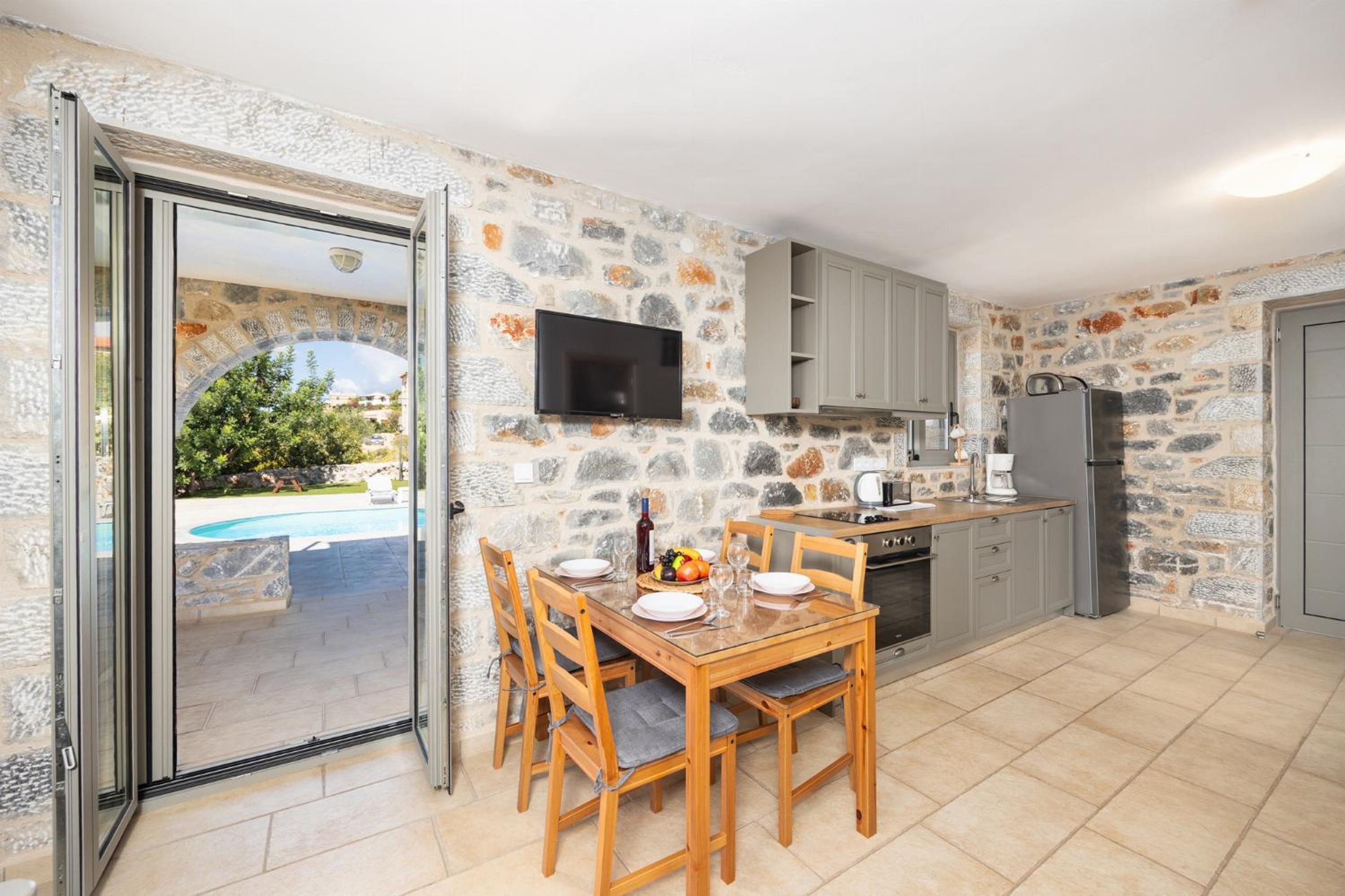 Una cuina amb parets de pedra, una taula de dinar amb caires, una televisió penjada sobre la barra i vista a una piscina a través d'una porta de vidre.