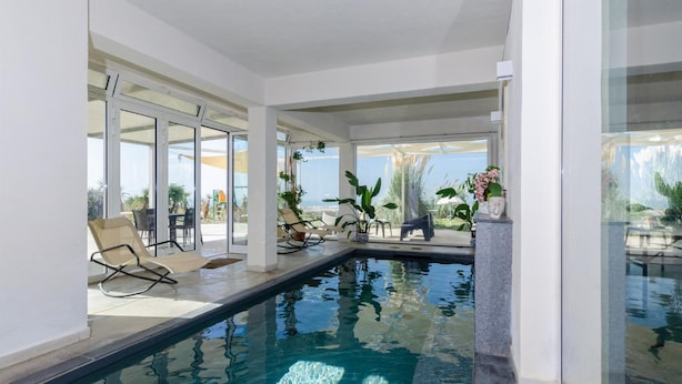 An indoor swimming pool with clear glass walls, lounge chairs, and potted plants. - Villa Padima in Scicli (Scicli)