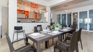 A modern dining area with a long table set for six, surrounded by wicker chairs. A kitchen with wooden cabinets and a built-in wine rack is visible in the background. - Villa Padima in Scicli (Scicli)