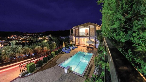 Et moderne hus med swimmingpool, en overdækket terrasse og udsigt over byen om natten.
