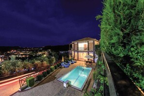 Een modern huis met een zwembad, een overdekt terras en uitzicht op de stad 's nachts.