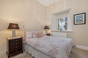A bedroom with a bed, two bedside tables, a dresser, a window with a view, and a framed picture on the wall. - Bryn Y Mor - Tenby Apartment Sea Views (Tenby)