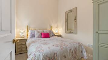 A neatly made bed with floral bedding, a bedside table with a lamp, and a wall-mounted mirror.