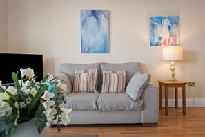 Ein Wohnzimmer mit grauem Sofa, gestreiften Kissen, einer Blumenvase, einer Lampe und zwei abstrakten Bildern an der Wand.