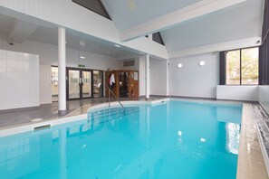 An indoor swimming pool with clear water, surrounded by white walls and columns, and a view of the outside through a large window. - Bryn Y Mor - Tenby Apartment Sea Views (Tenby)