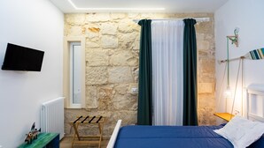 Une chambre avec un mur en pierre, un lit avec une literie bleue, un banc en bois, une télévision et une fenêtre avec des rideaux.