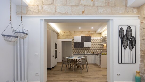Une cuisine moderne avec un mur en pierres apparentes derrière le plan de travail, un coin repas avec une table ronde et des chaises, et un panier suspendu.