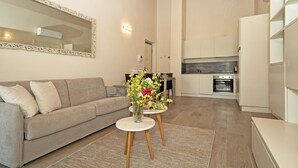 Una sala de estar con un sofá, una mesa de centro con un jarrón de flores, una cocina al fondo y un espejo grande en la pared.